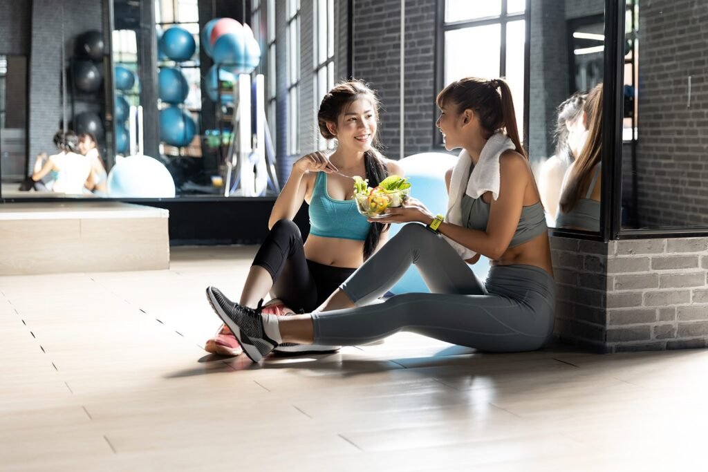 Two women sitting in a gym, sharing a nutritious diet bowl, as they embark on their 'How to Lose Belly Fat Zero Belly Diet' adventure together.