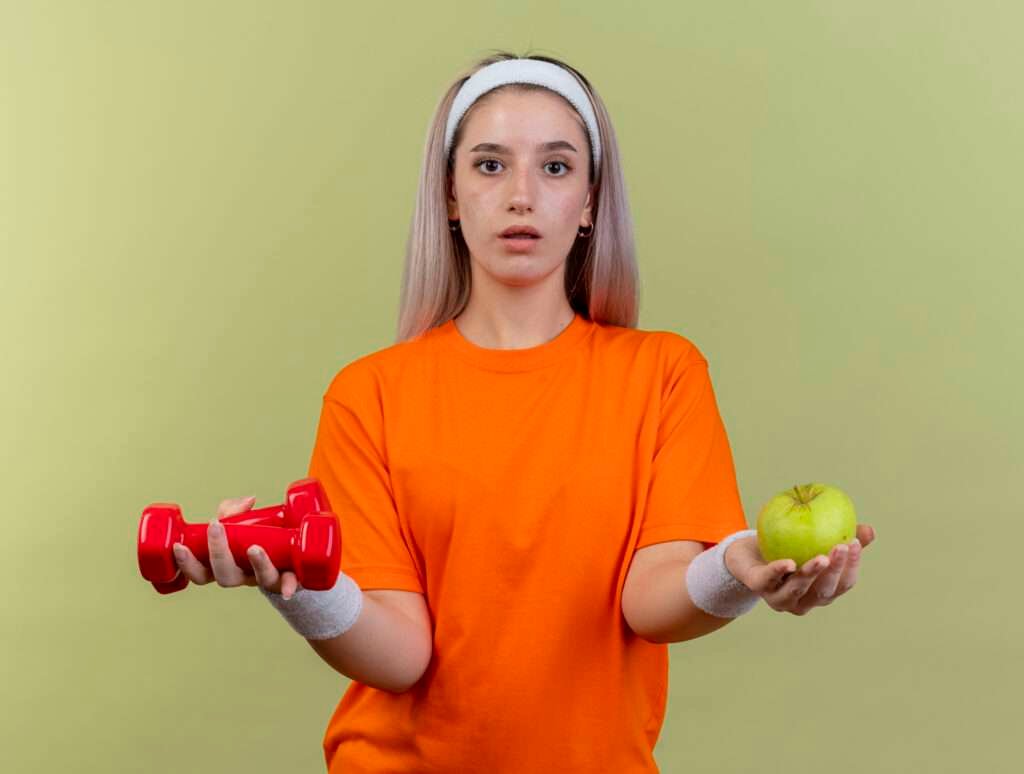 Young Caucasian sporty girl with braces, headband, and wristbands holding dumbbells and an apple, embodying the commitment to health and fitness while delving into the fundamentals of weight loss.