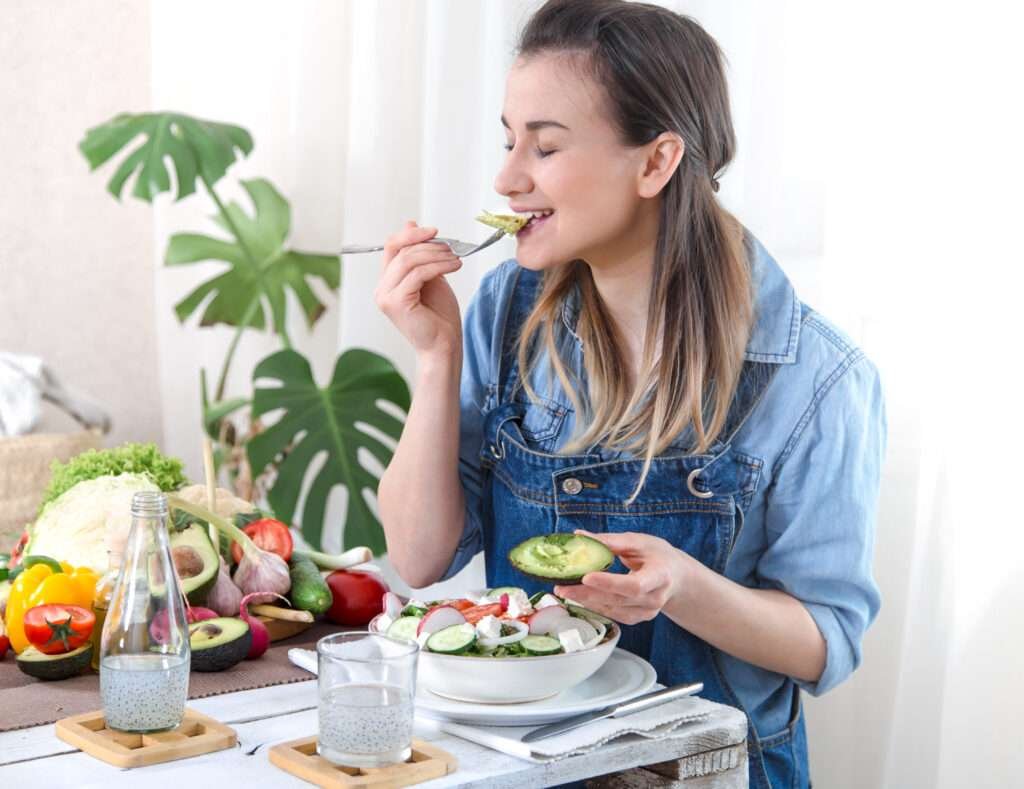 A young and joyful woman in denim attire relishing a salad aligned with her Keto lifestyle, made with organic vegetables at the table, set against a light background. Illustrating the concept of wholesome homemade nutrition and well-being.